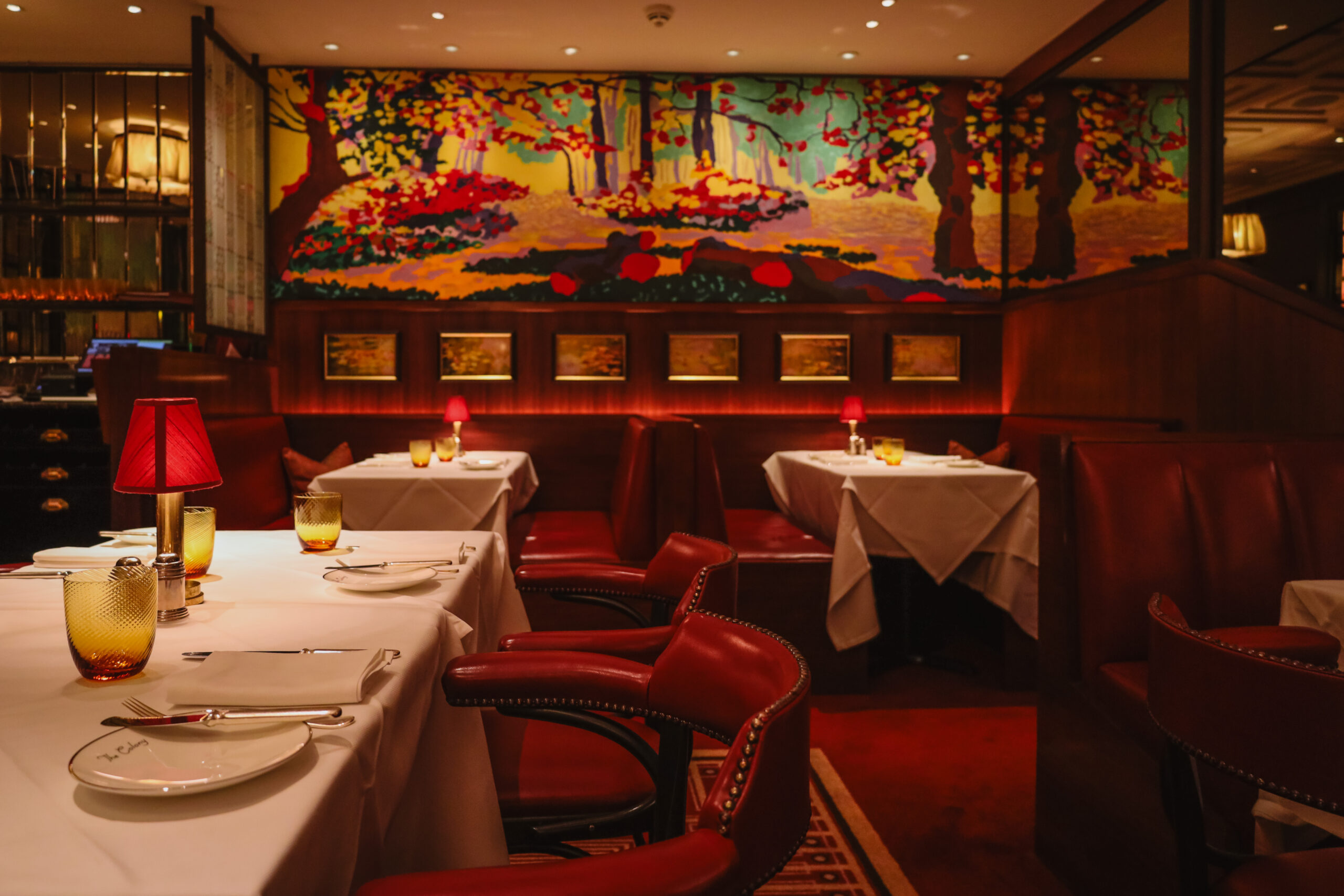 Red banquettes and white table cloths in dim, steakhouse-style lighting with a colorful watercolor mural in the background of this restaurant dining room.