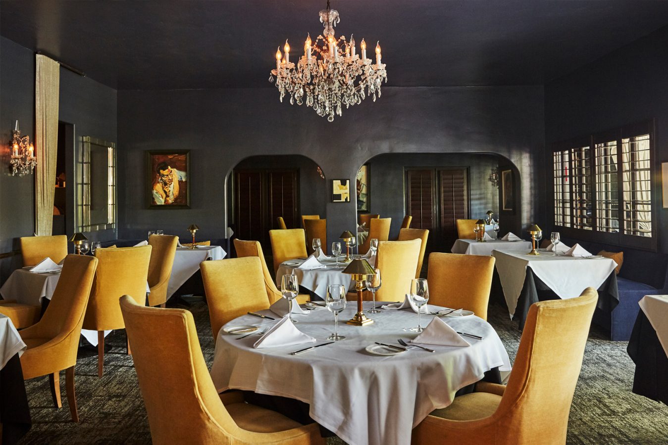 A grand dining room with yellow velvet chairs, white table cloths, a lit chandelier and dark walls in this upscale restaurant.