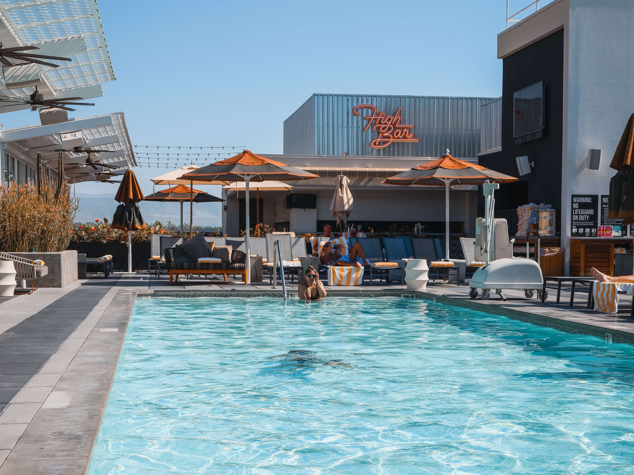 A view of the rooftop pool from sitting poolside with someone swimming in the foreground. The High Bar and umbrella covered tables feature in the background.