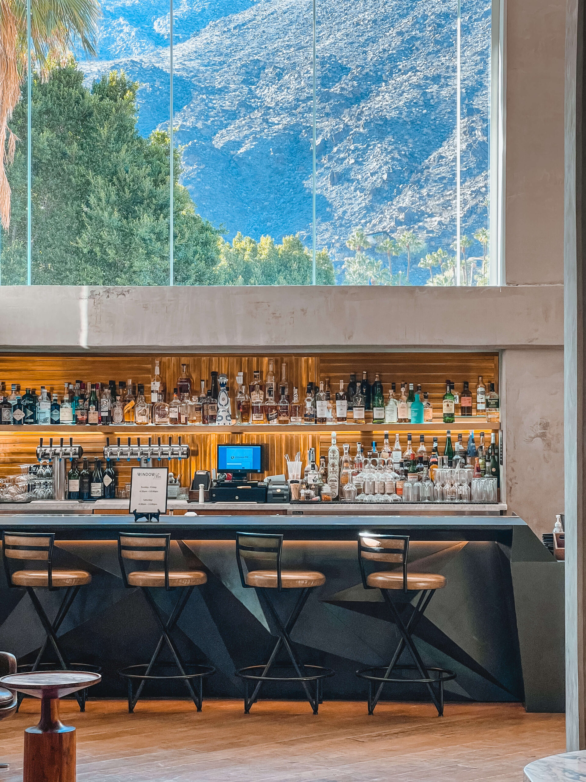 View of the lobby bar at the Kimpton hotel with four bar stools in front of a bar and a large window overhead framing the view of the San Jacinto Mountains in the background.