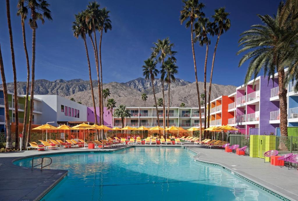 A view from the pool at the Saguaro hotel with brightly painted rooms lining around the pool area. Palm trees and the mountains behind Palm Springs can be seen in the background.